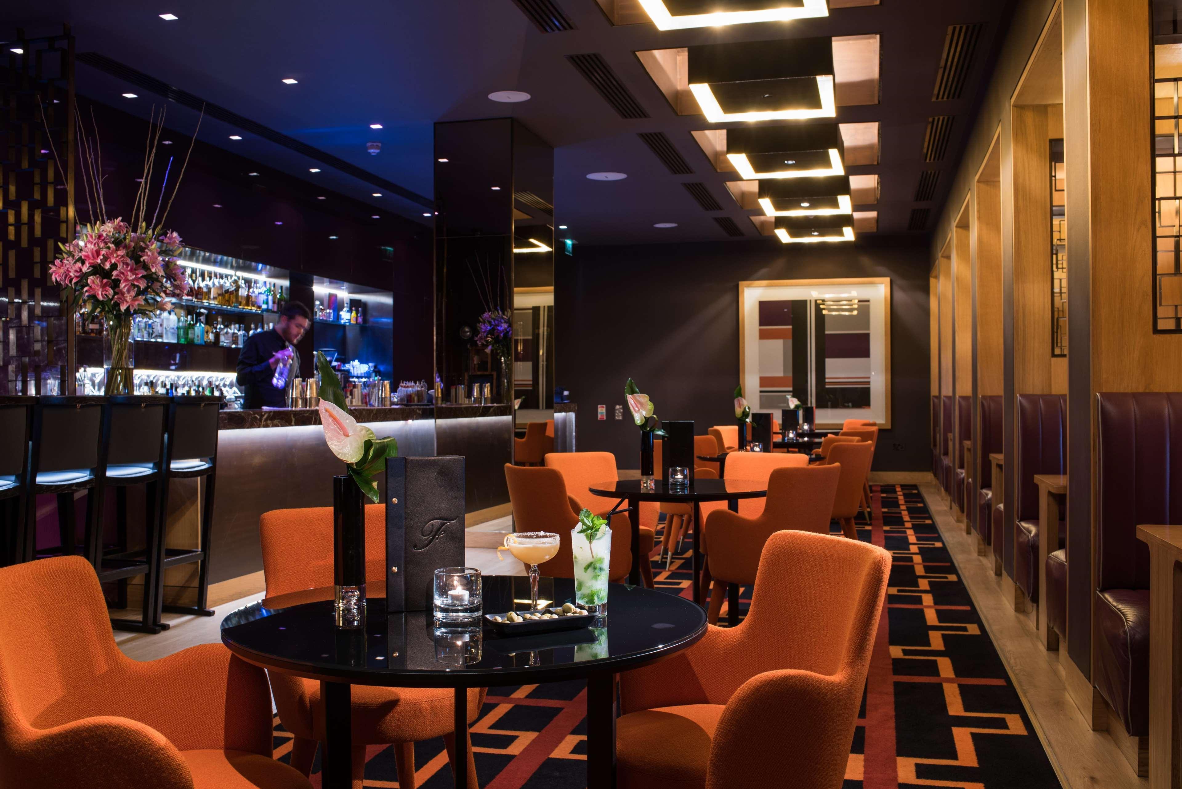 The Fitzwilliam Hotel Belfast Eksteriør bilde The photo depicts a stylish bar or lounge area with a modern and sophisticated atmosphere. The space features dark walls accented by blue lighting and a sleek bar counter in the background, stocked with various drinks. The seating consists of round t