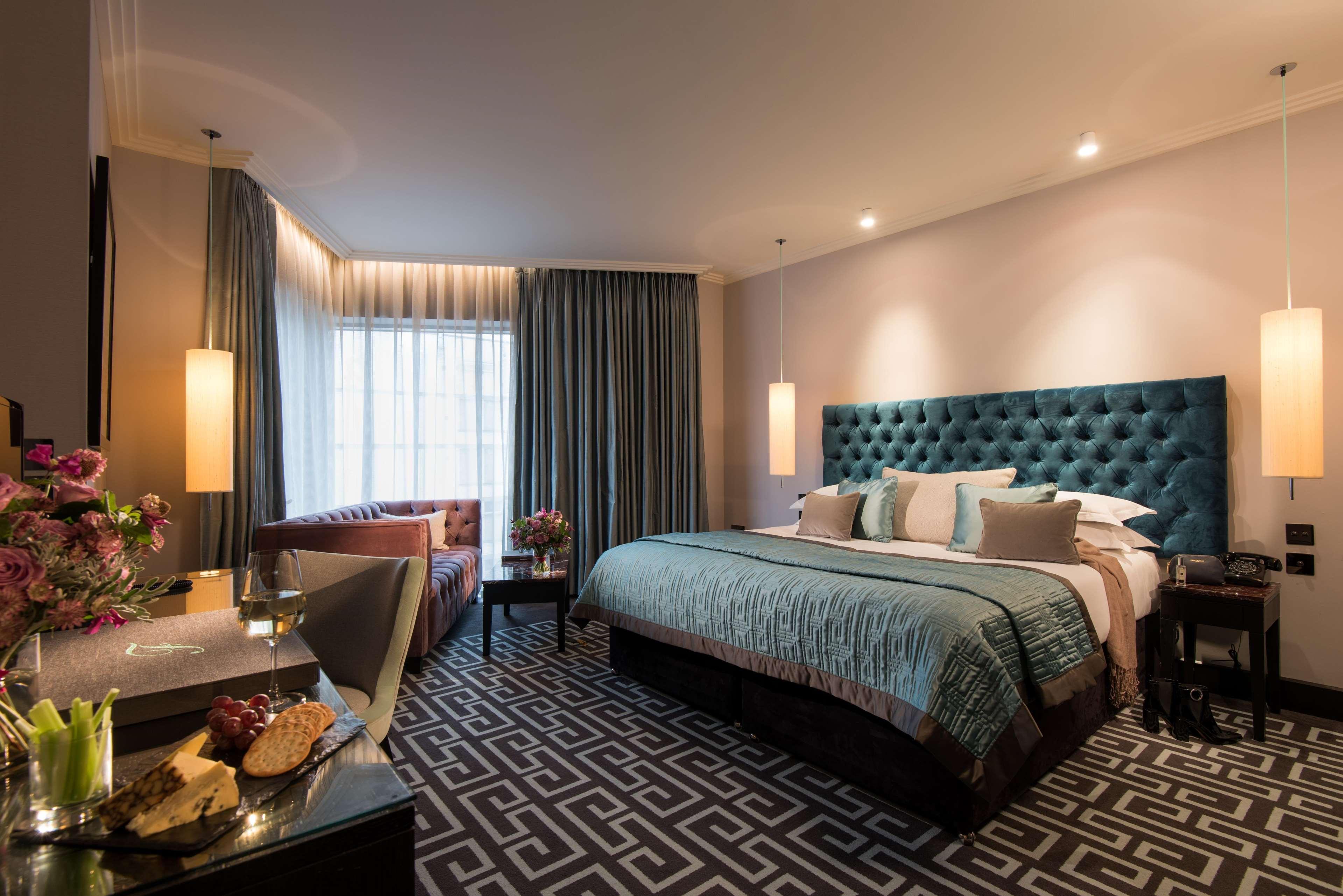 The Fitzwilliam Hotel Belfast Eksteriør bilde The photo depicts a stylish hotel room. It features a large bed with a tufted teal headboard, flanked by two wall sconces providing soft lighting. The bedding is neatly arranged with accent pillows. To one side, there is a small seating area with a c