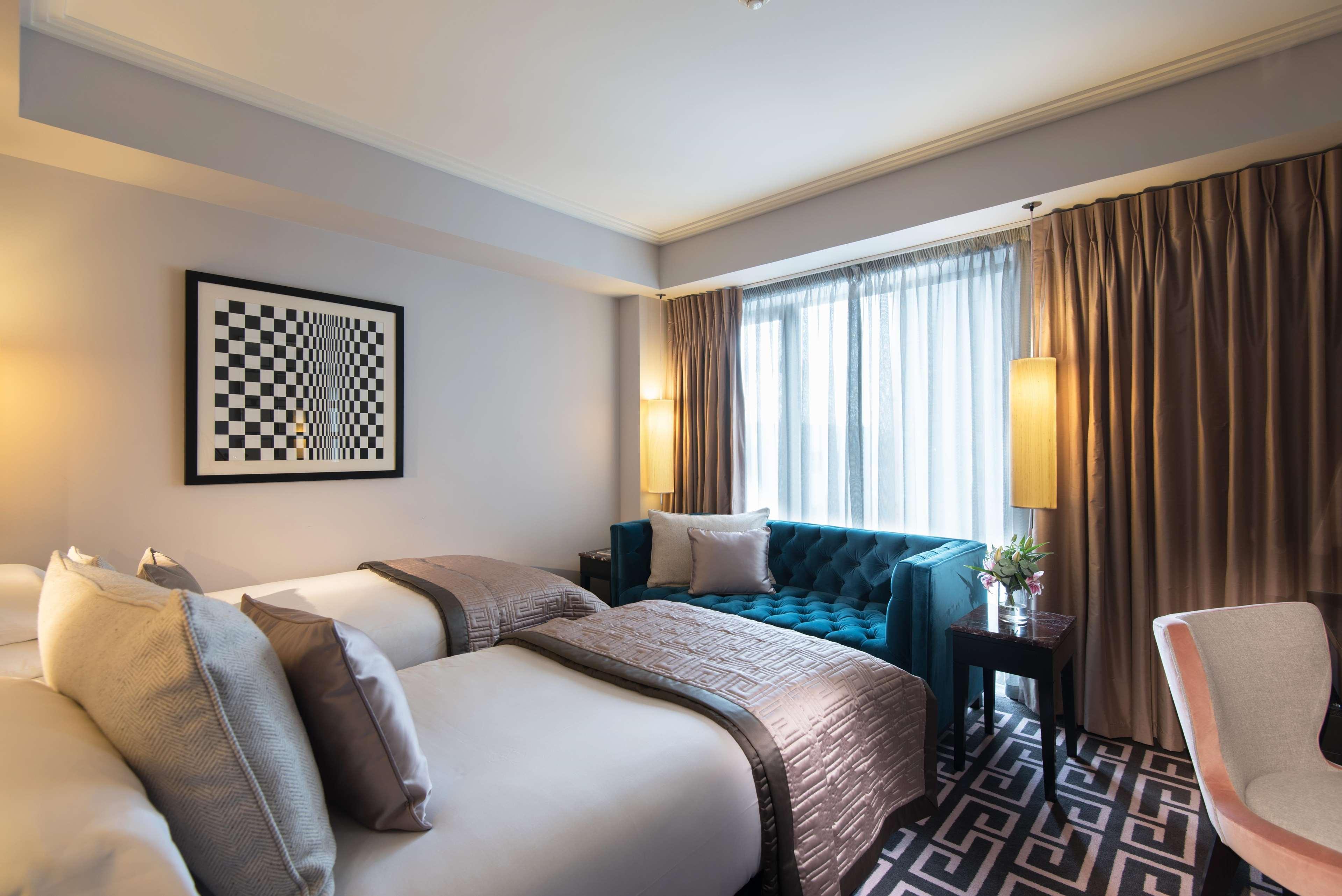 The Fitzwilliam Hotel Belfast Eksteriør bilde The photo shows a modern hotel room. It features two beds with decorative pillows, a stylish teal couch, and large windows with sheer curtains allowing natural light to enter. The walls are painted in neutral tones, and there is a piece of artwork wi