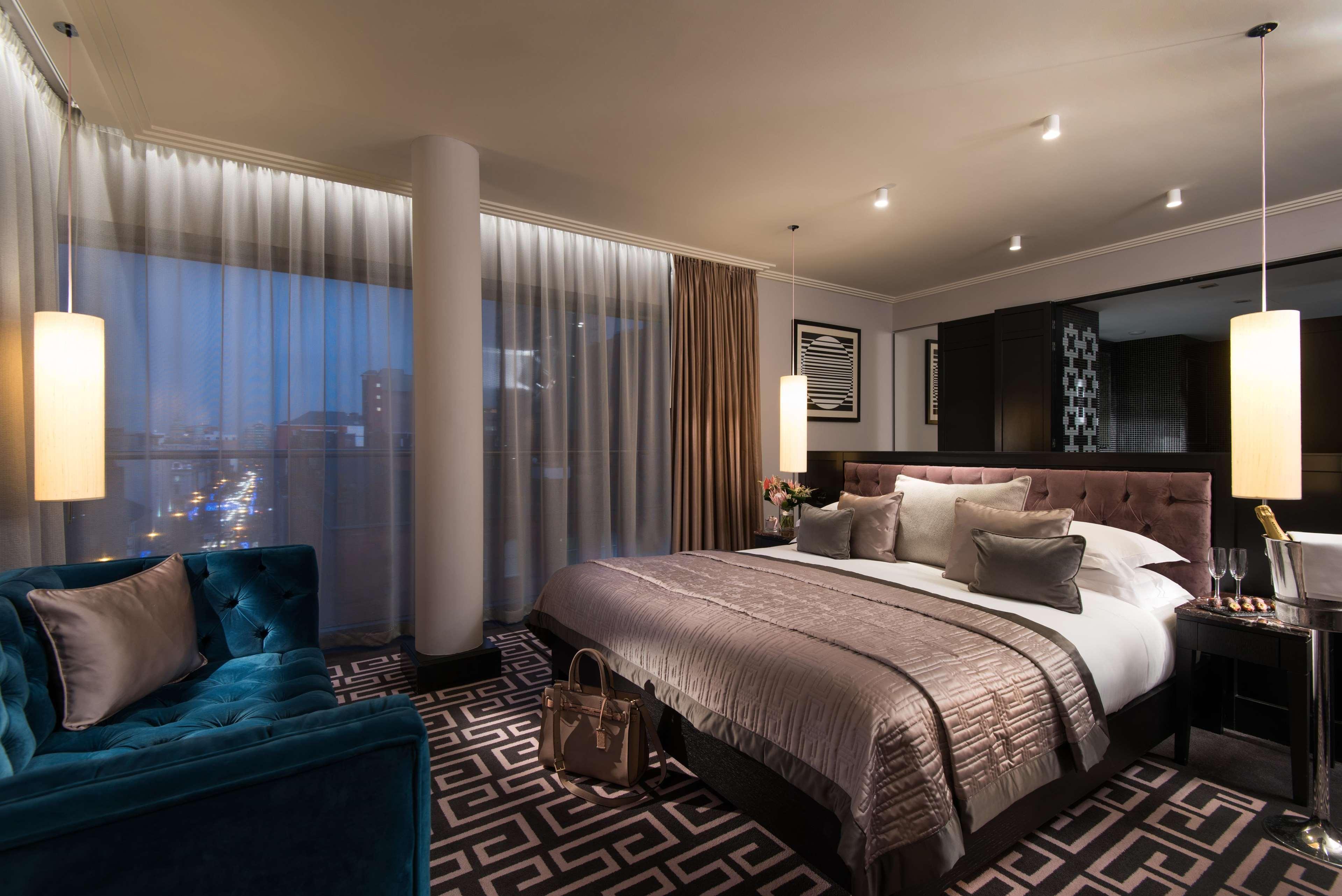 The Fitzwilliam Hotel Belfast Eksteriør bilde The photo shows a luxurious hotel room with a modern design. It features a large bed with decorative pillows and a stylish bedspread, complemented by bedside lamps. To one side, there is a plush turquoise sofa and a coffee table. The room has floor-t