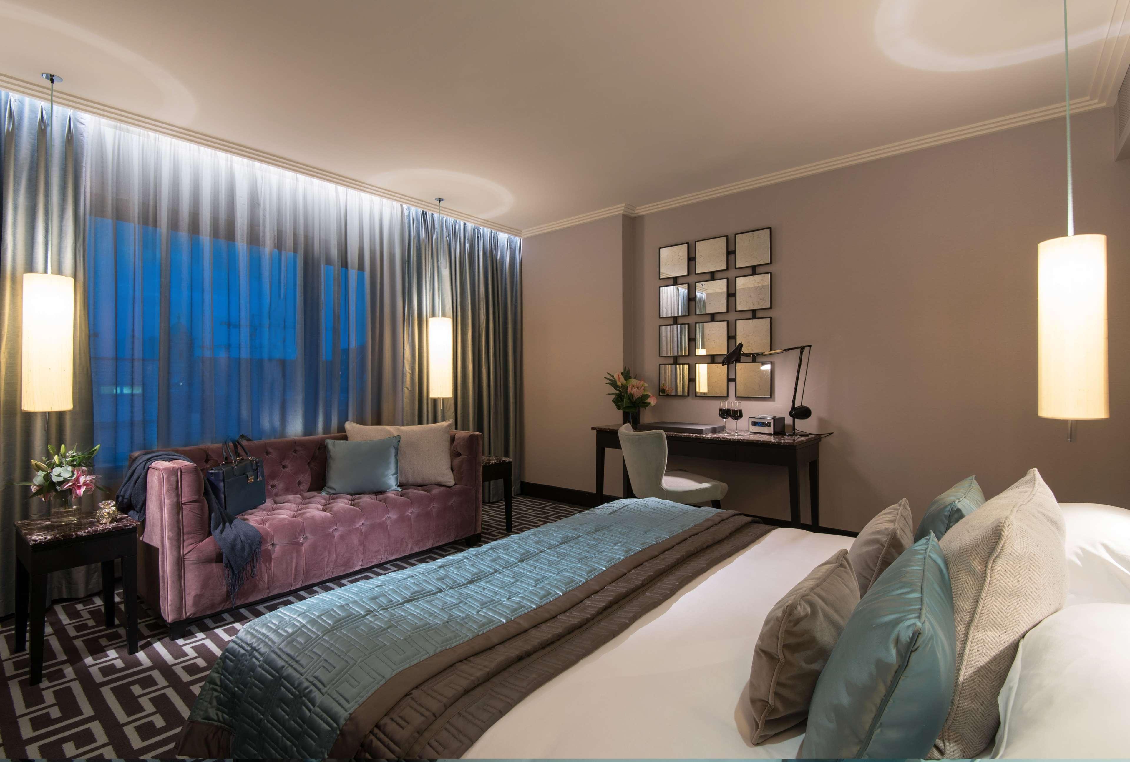 The Fitzwilliam Hotel Belfast Eksteriør bilde The photo depicts a modern hotel room featuring a spacious layout. There is a large bed with decorative pillows in the foreground, covered with a stylish bedspread. To one side, a plush purple sofa adds a pop of color and comfort. A small desk and ch