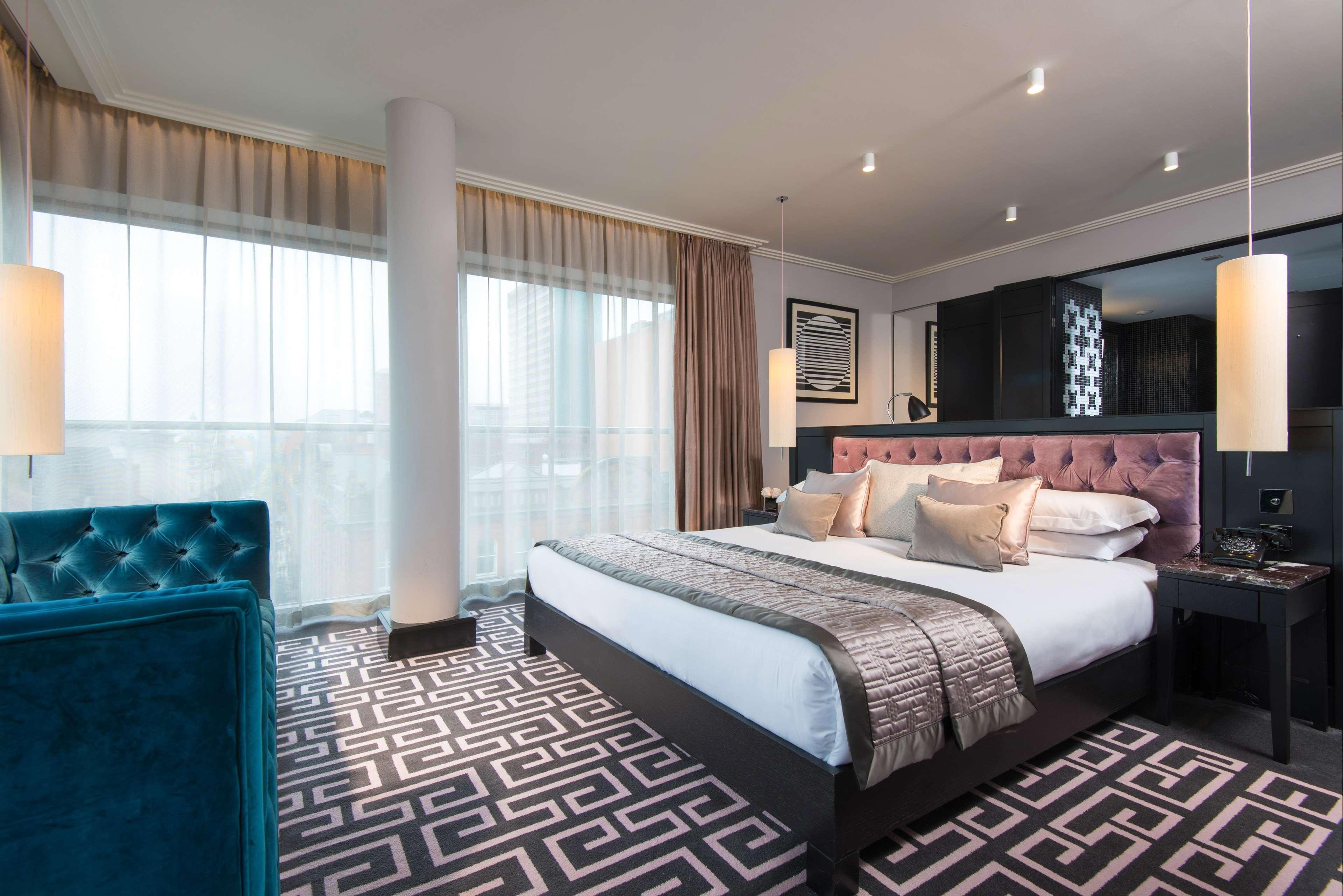 The Fitzwilliam Hotel Belfast Eksteriør bilde The photo showcases a modern hotel room. It features a large bed with plush pillows and a stylish headboard. The room has large windows with sheer curtains, allowing natural light to filter in. The floor is covered with a patterned carpet that contra